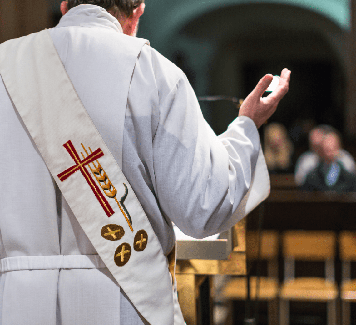 A priest is wearing a white robe and holding his hand up.